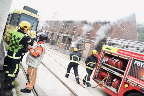 Image of recruit fire fighters with a fire engine and burning building in the background.